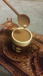 A spoon, covered in thick drinking chocolate, hovers above a bronze Turkish coffee cup.