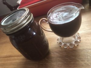 A small, clear glass pitcher filled with hot fudge. Behind it is a large canning jar fill to the brim with hot fudge!