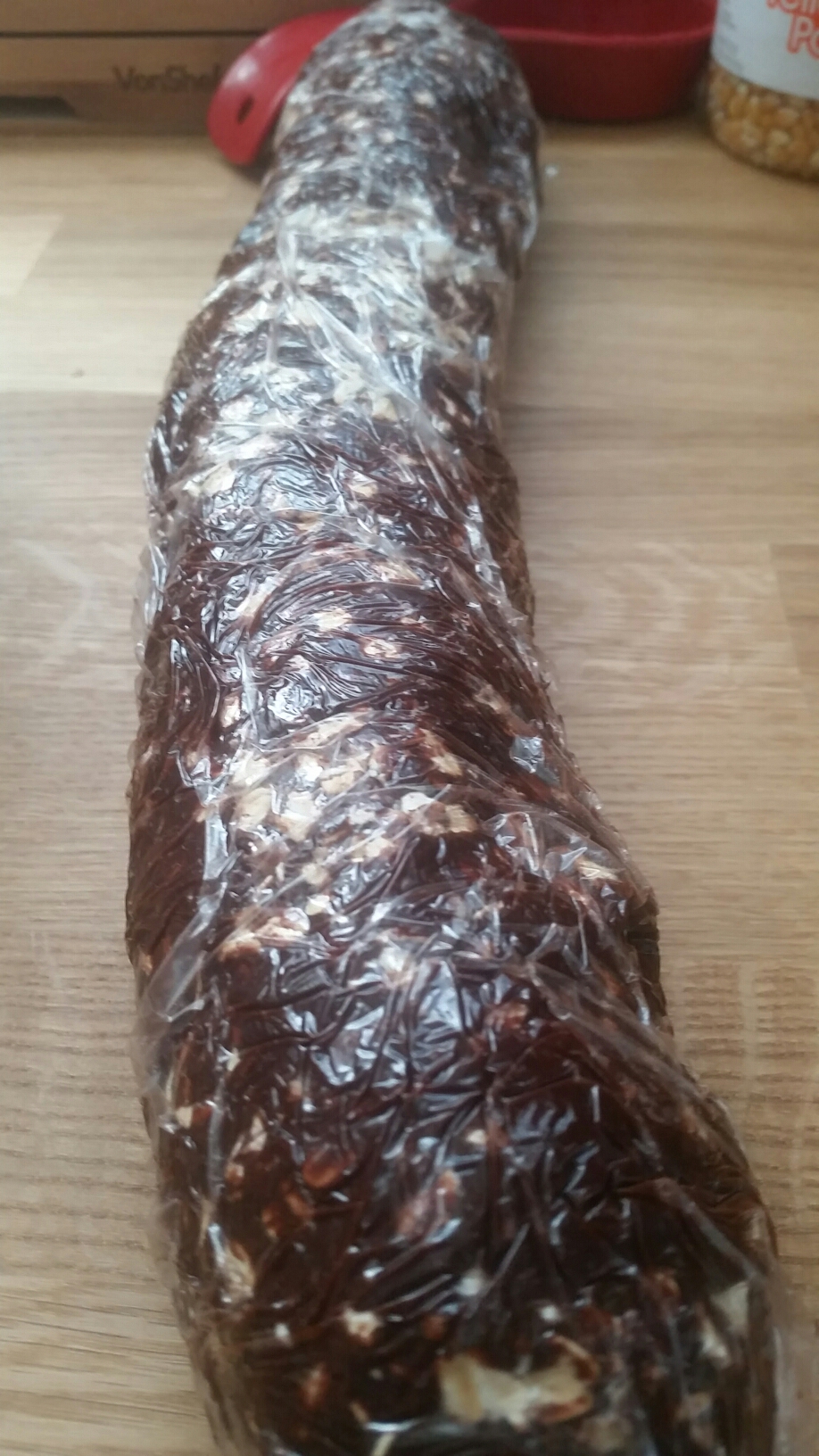 A cling wrapped log of brown cookie dough with flecks of white embedded in it.