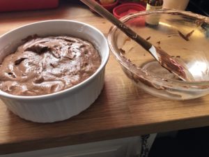 A baking dish is filled with chocolate souffle batter sits next to an empty mixing bowl with chocolate streaks still in it.